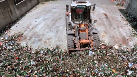 la excavadora de carretilla está agarrando vidrio y cargándolo en la parte trasera de un camión transportador, la máquina de servicio pesado tiene un brazo largo equipado con un accesorio para agarrar vidrio, vista de drones, concepto de reciclaje de vidrio