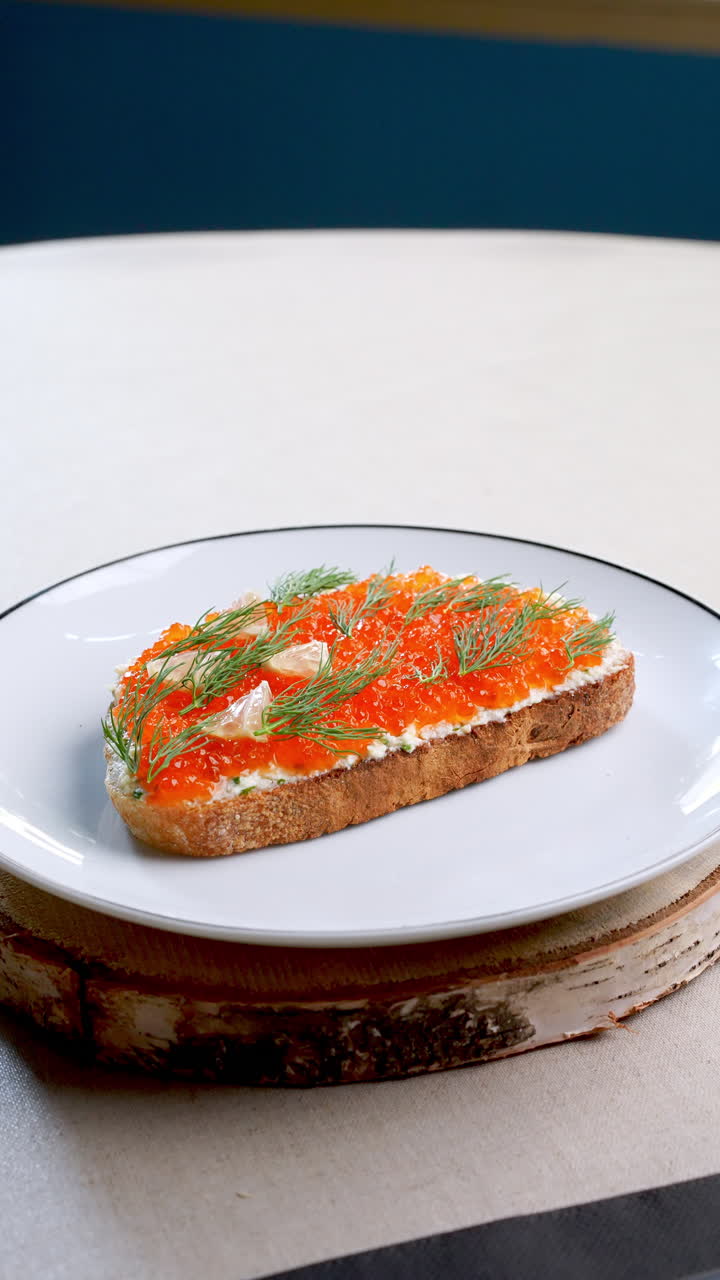 Gourmet appetizer featuring cream cheese topped toast decorated with vibrant red caviar and fresh dill, elegantly plated on pristine white dish with dark rim resting atop rustic birch wood slice