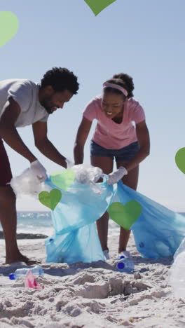 animación de corazones verdes sobre una pareja afroamericana con bolsas de reciclaje limpiando la playa