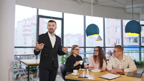 businessman giving a talk to the camera and gesturing with his hands. team of workmates sit next at the table having a conversation.