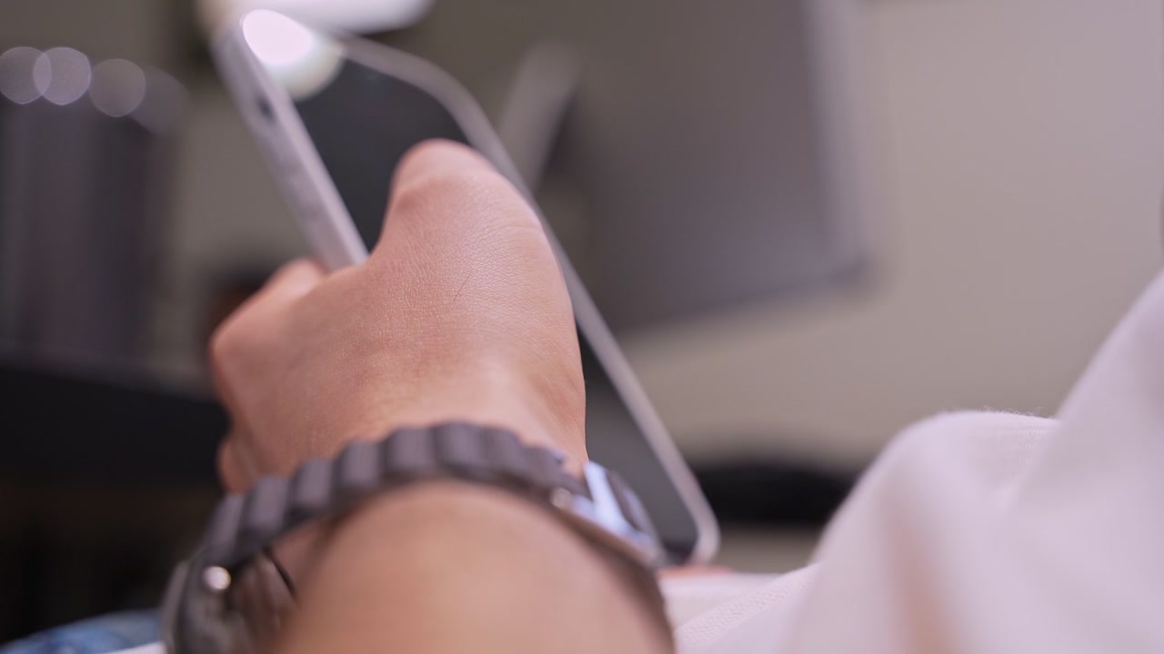Primo piano della mano di una persona che indossa uno smartwatch, che tiene in mano e naviga su uno smartphone. Scatto con profondità di campo ridotta in un moderno ufficio domestico con un monitor di computer sfocato sullo sfondo