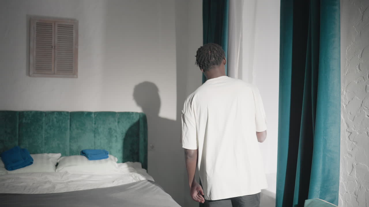 Person in white shirt standing near window, adjusting curtain, soft natural light streaming through, peaceful bedroom with green velvet headboard and fresh linens, modern minimalist decor