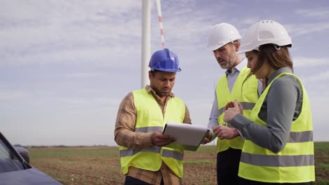 three caucasian and latin engineers standing on wind turbine field and discussing over plastic models.