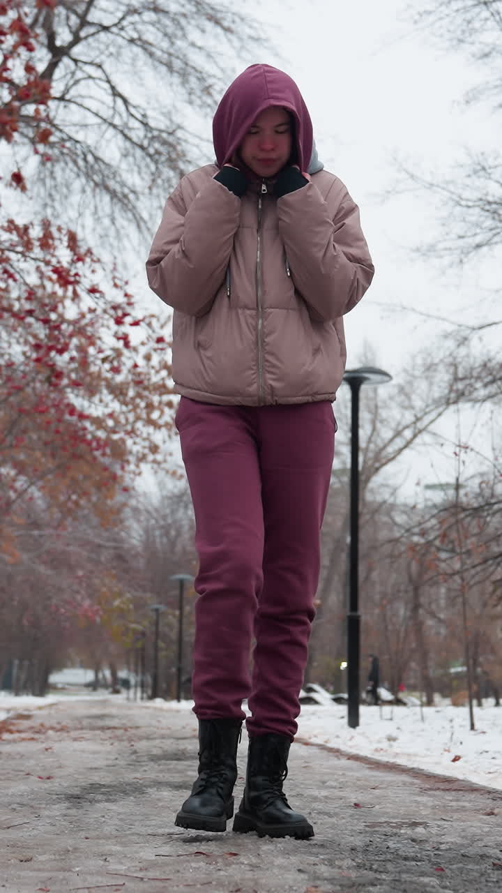 mujer en traje de invierno paseando sola por un parque cubierto de nieve, ajustando la capucha mientras trae la mano cerca de la boca para mantener el calor, árboles desnudos, nieve esparcida y cielo nublado