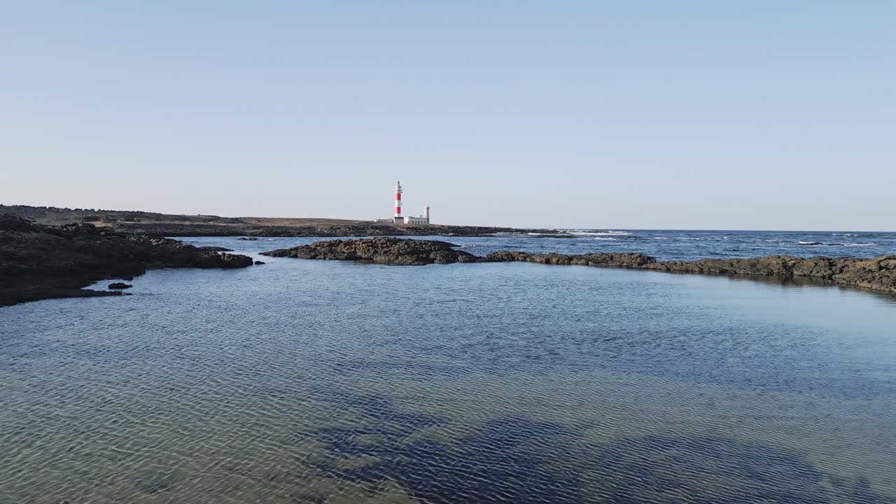 Aerial drone shot flying toward El Tostón Lighthouse in El Cotillo, Fuerteventura, with volcanic coastline and Atlantic waves in view.