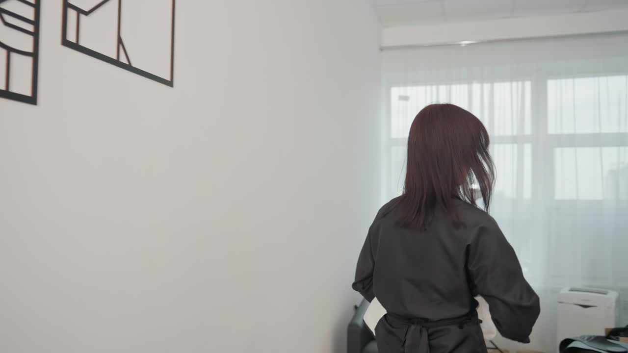 white brunette stylist celebrates in salon, fist pump after successful consultation, walks to shelf, reaches products, greets blonde client seated in chair near mirror and natural light