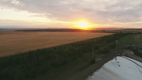 Hochwertige-Aufnahmen-Einer-Drohne,-Die-Bei-Sonnenuntergang-Von-Feldern-In-Der-Natur-Wegfliegt-Und-Eine-Geomembran-Freilegt,-Die-Den-Kontrast-Zwischen-Natürlichen-Landschaften-Und-Mülldeponien-Verdeutlicht