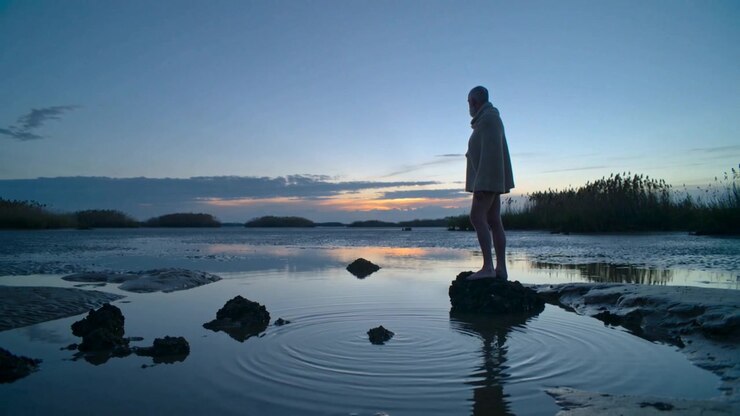 Standing senior man in cloak scanning horizon at tidal flat, stone ripples urging look, copy space