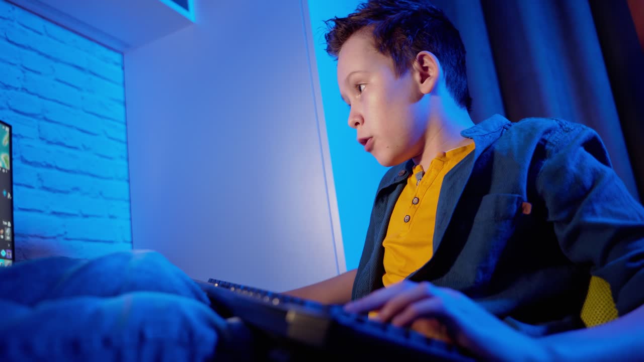 Excited boy playing and winning in online video game. Teenager sitting comfortably at home and playing on his personal computer. Blue light in the room.