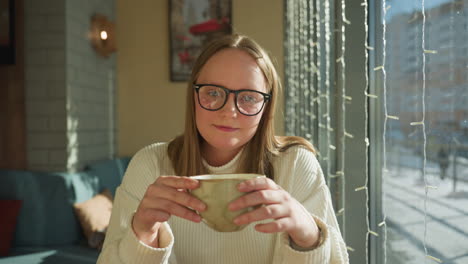 close up portrait of young woman in glasses holding ceramic mug while sitting near large window with hanging decorative lights, as sunlight brightens cozy indoor scene with soft reflections