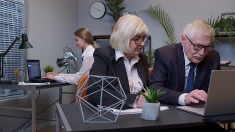 elderly man boss with woman colleague working in office, developing new project on laptop, team work