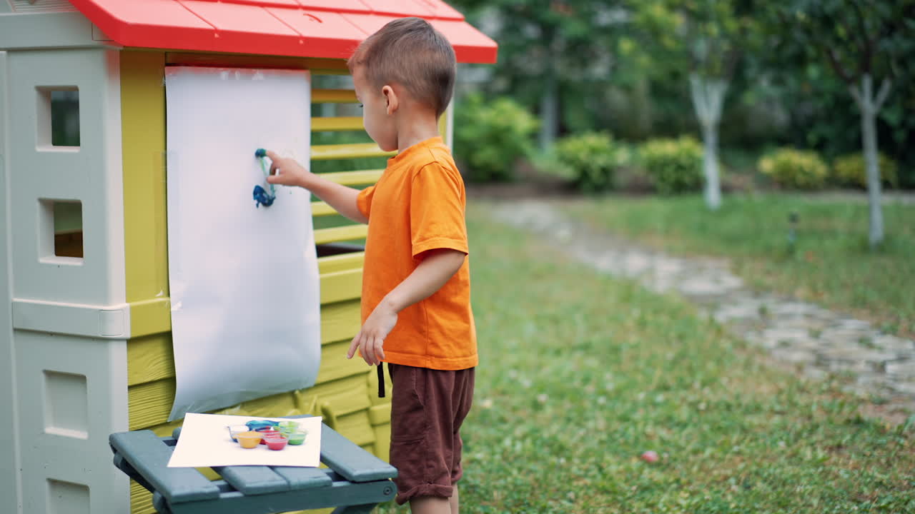 Toddler kid in orange t-shirt and brown shorts takes some paint with his finger. Creative baby boy painting outdoors.