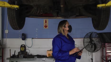 mecánico femenino con máscara facial sosteniendo el clipboard e inspeccionando el coche en una estación de servicio de coches
