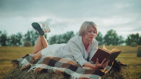 focus on woman with leg raised on hay bale, checkered blanket, reading book, strand of hay in mouth, sun hat beside her hand, enjoying peaceful reading time in an open field, flipping pages