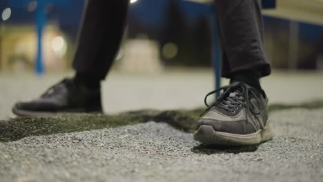 close-up of a person's legs, focusing on their shoes as he continuously tap their foot on the ground, the background is blurred and dimly lit, with shadows visible on the ground