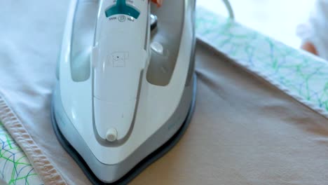a steam iron glides over beige trousers on a patterned ironing board, releasing bursts of steam in bright, natural daylight. camera remains steady, focusing on the ironing process