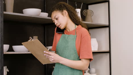 young female clerk having a call and writing on a clipboard in the pottery shop