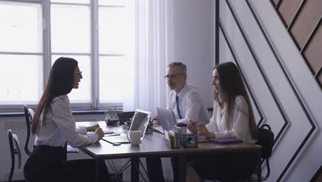 one male co-worker and two female co-workers talking between them in front of laptops at the office