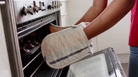 Midsection-of-unaltered-african-american-woman-taking-cakes-out-of-oven-in-kitchen,-in-slow-motion