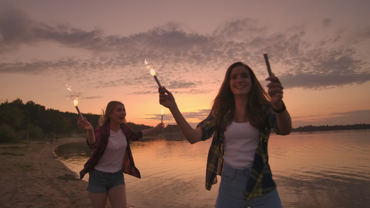 felices chicas sonrientes corriendo por la playa con chispas por la noche. sostengan las luces brillantes y rían estudiantes jóvenes despreocupados