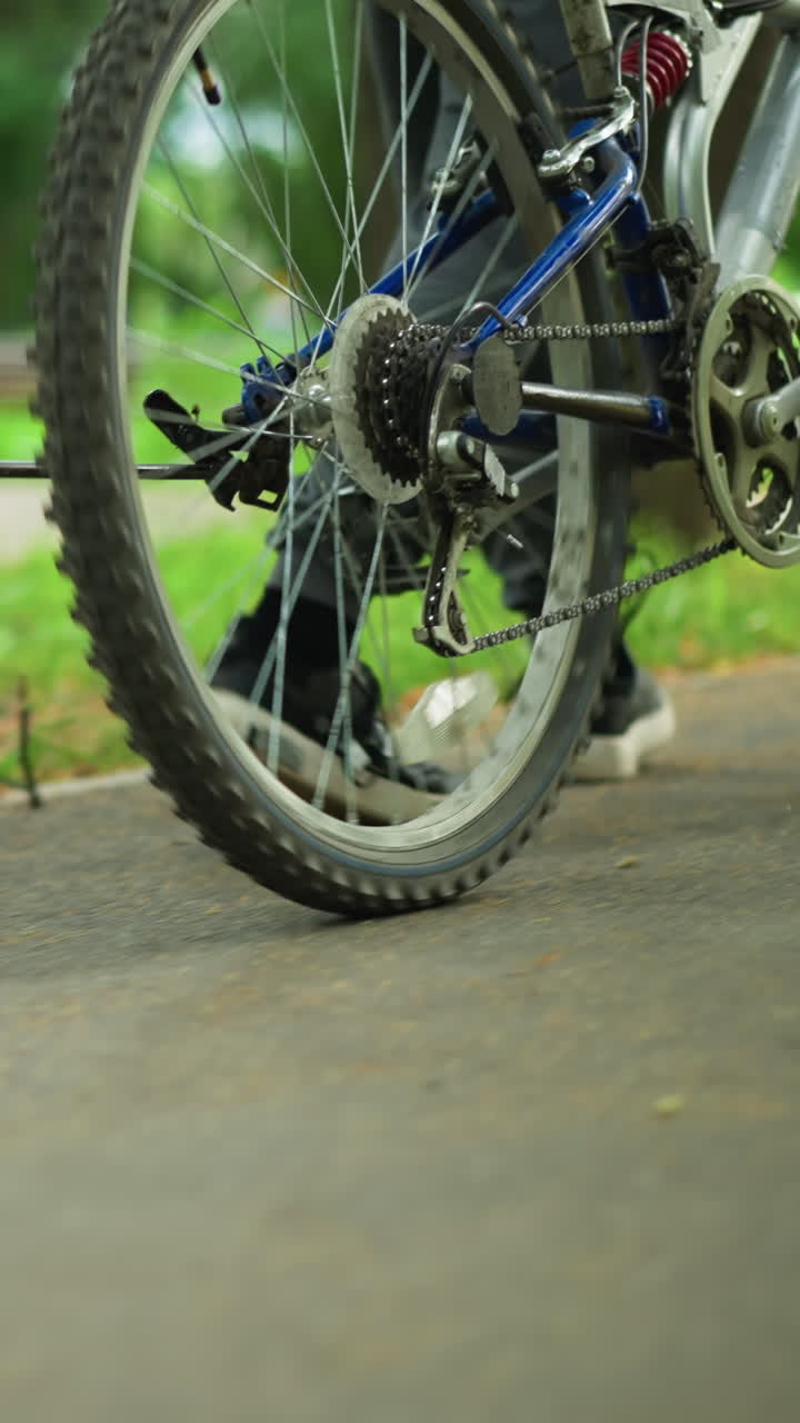 Close-up of someone walking beside bicycle along a park walkway, with grassy background, the shoelaces float as he move, the focus is on the legs and bike, with trees in the background