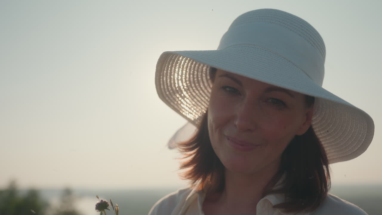 elegant woman in white hat smiles gently while holding bouquet of wildflowers under warm sunlight, serene nature background with soft sky glow enhances peaceful countryside afternoon moment