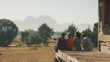 Rear-View-Of-Friends-On-Vacation-Sitting-On-Porch-Of-Countryside-Cabin-Taking-Selfie-On-Mobile-Phone