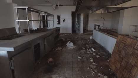 abandoned industrial kitchen with trash on the floor and a decaying ceiling.  static shot showing the cabinets, countertops, shelves, and ceiling vents.
