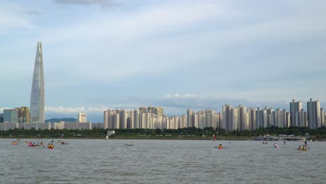 Menschen-Gruppieren-Kajakfahren-Und-Windsurfen-Am-Fluss-Han-Mit-Dem-Berühmten-Lotto-turm-Von-Seoul-Im-Hintergrund-Bei-Sonnenuntergang