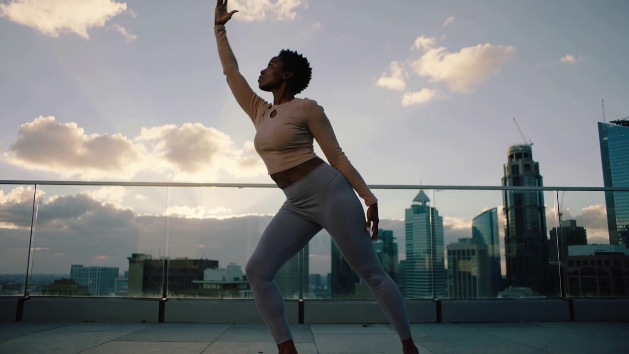 Lifting arm, woman in grey leggings beige top practicing balance on rooftop with glass balustrade