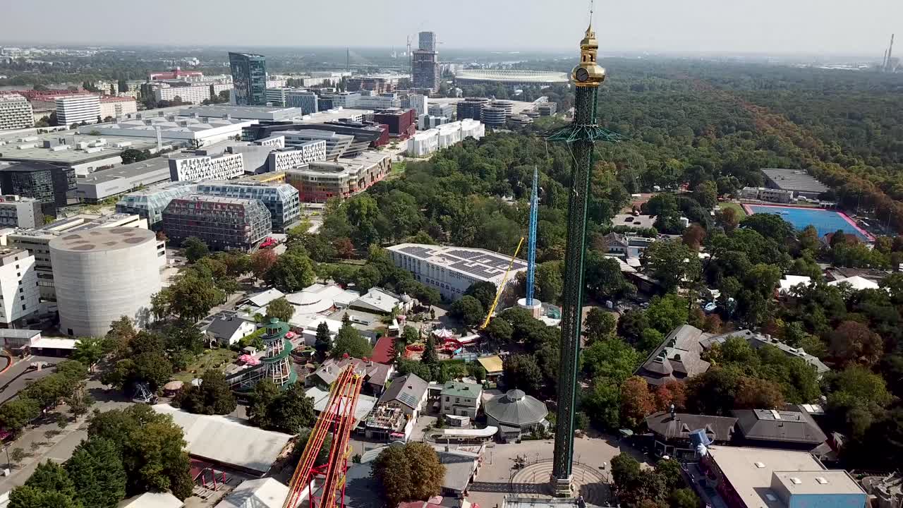 Vienna Prater with Tower in amusement park during sunset. Cityscape view with downtown skyscrapers in backdrop. Aerial orbit wide shot.