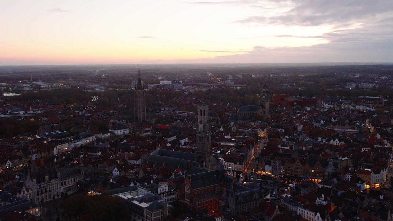 Riprese con drone che mostrano un'ampia panoramica di Bruges di sera, con il paesaggio urbano storico, edifici illuminati e un cielo parzialmente nuvoloso.