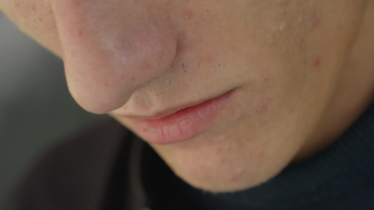 Primo piano di labbra e mento di un uomo caucasico, luce interna soffusa, dolcevita, espressione pensosa e delicata, attore che prova le battute, che finge di parlare, acne leggera e barba incolta visibili, cinematografico e intimo
