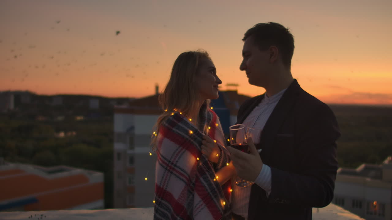 젊은 부부는 지붕에서 포옹하고 컵에서 은 와인을 마시며 색 옷을 입고 서서 도시 위의 아름다운 일몰을 감상하고 있습니다.