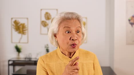 elderly asian woman in yellow top makes a quiet gesture with finger to lips, in a softly lit, modern home interior with framed art