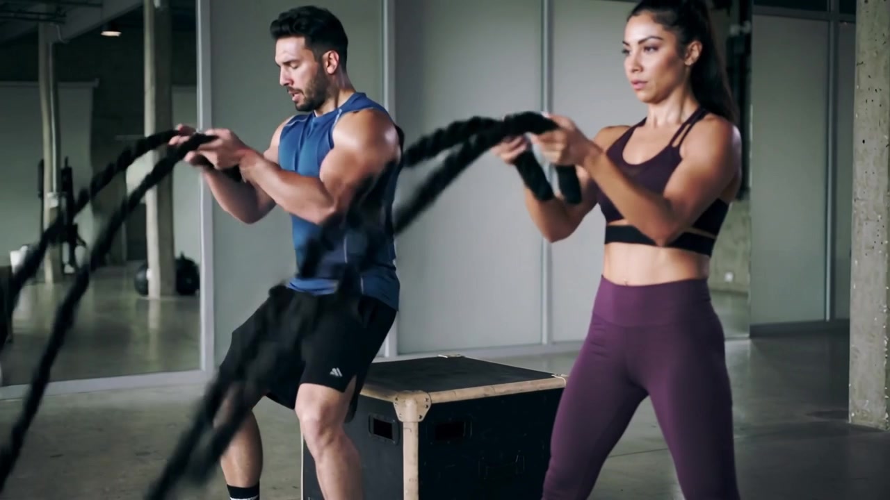 Hombre con camiseta azul sobre cajón pliométrico y mujer con leggings morados, agarrando cuerdas y alternando ondas en un gimnasio de entrenamiento.