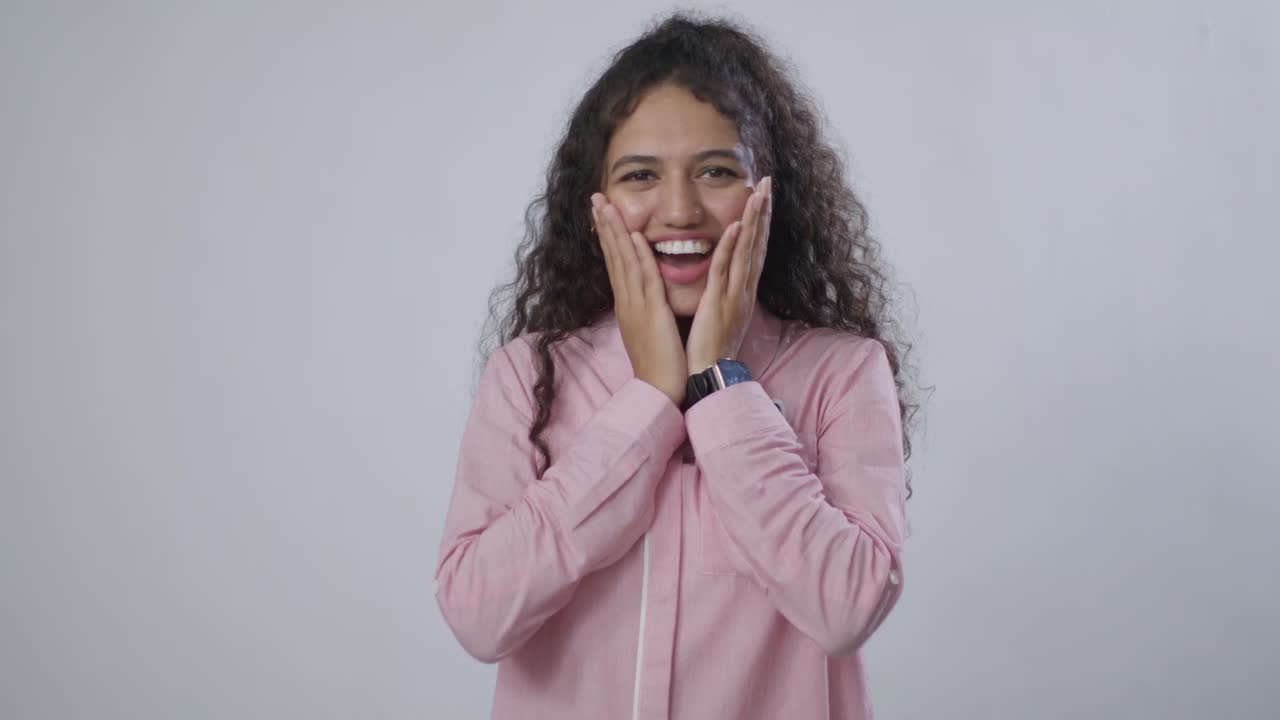 A South Asian woman with curly hair, wearing a pink shirt, looks surprised and happy, hands on cheeks