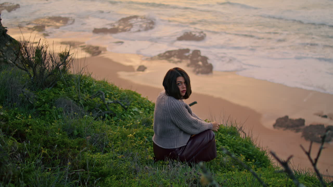 Sad woman sitting grass seacoast in front ocean autumn evening. Model posing.