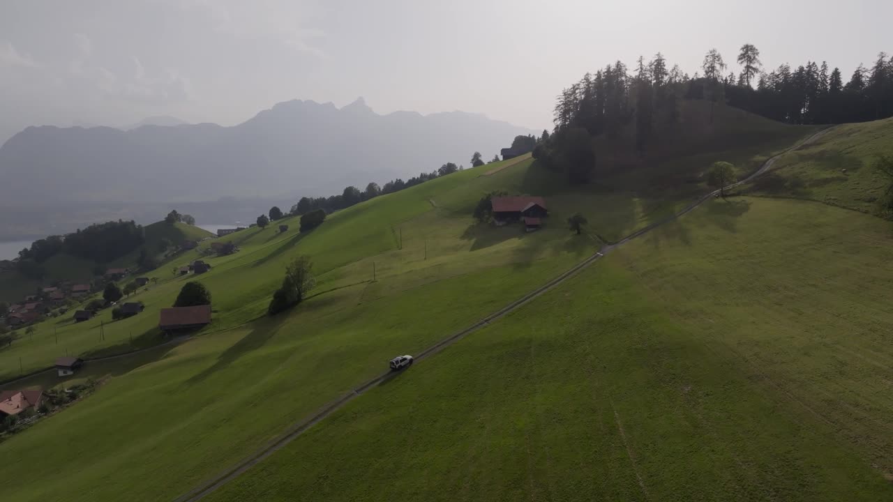 A white car drives along a narrow road winding through lush green alpine meadows, surrounded by scattered houses, trees, and distant misty mountains under soft daylight