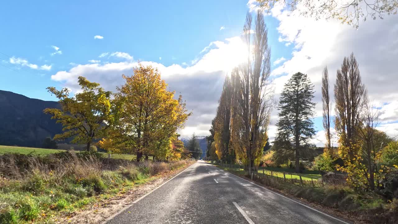 Timelapse of car driving along rural road, autumn trees, mountains, dramatic sunlight, wide angle