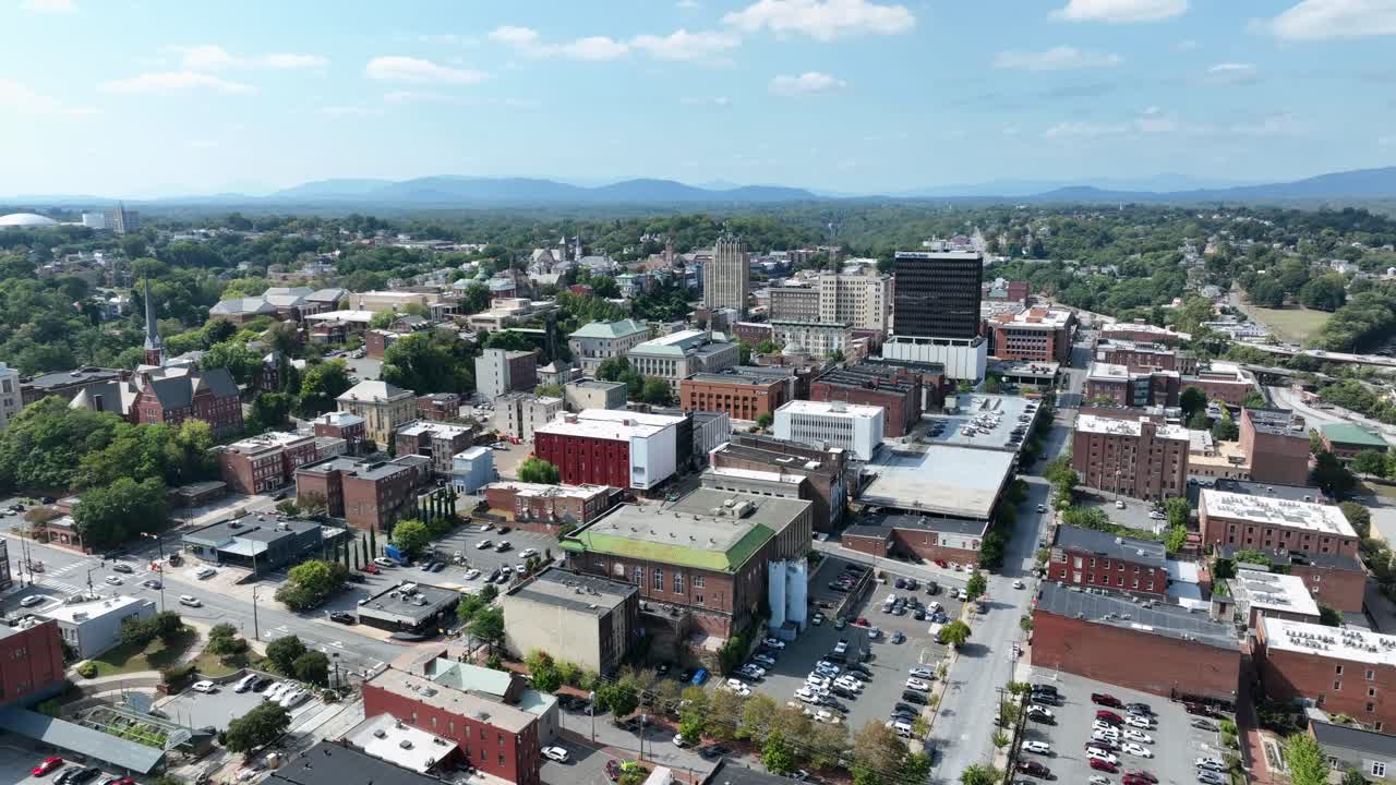 Downtown Lynchburg, Virginia, with historic architecture, modern buildings, surrounded by green hills and framed by distant Blue Ridge Mountains in vibrant cityscape. Aerial wide shot