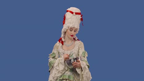 portrait of courtier lady in vintage lace dress and wig is preparing food, whisking something in a bowl. young woman posing in studio with blue screen background. close up. slow motion ready 59.94fps