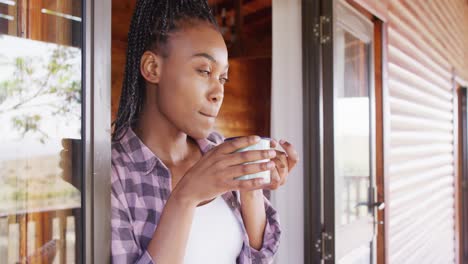 African-american-woman-looking-through-window-and-drinking-coffee-in-log-cabin,-slow-motion