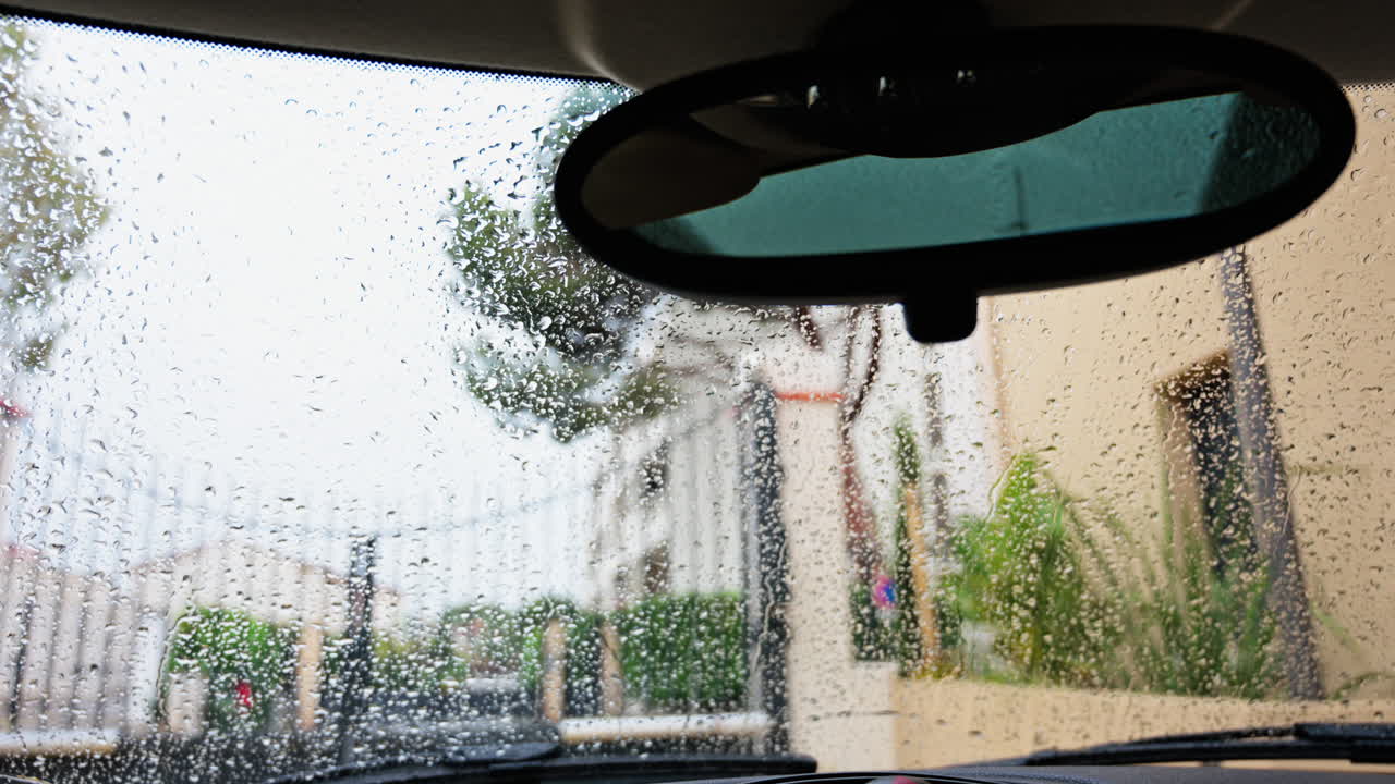 Close up of raindrops falling on the windshield of a car with a view of a gate, a fence and trees