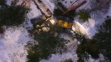 Wheeled-harvester-turns-on-snowy-ground-to-fell-tree-at-night,-top-down-aerial