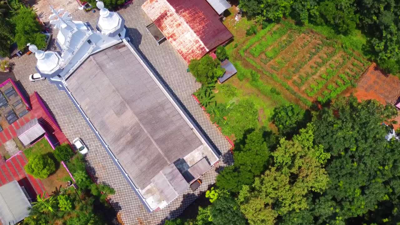 Aerial top-down view of a beautiful white church and a farm, surrounded by lush green tropical trees. Slow descent drone shot of a Christian religious building in a rural area
