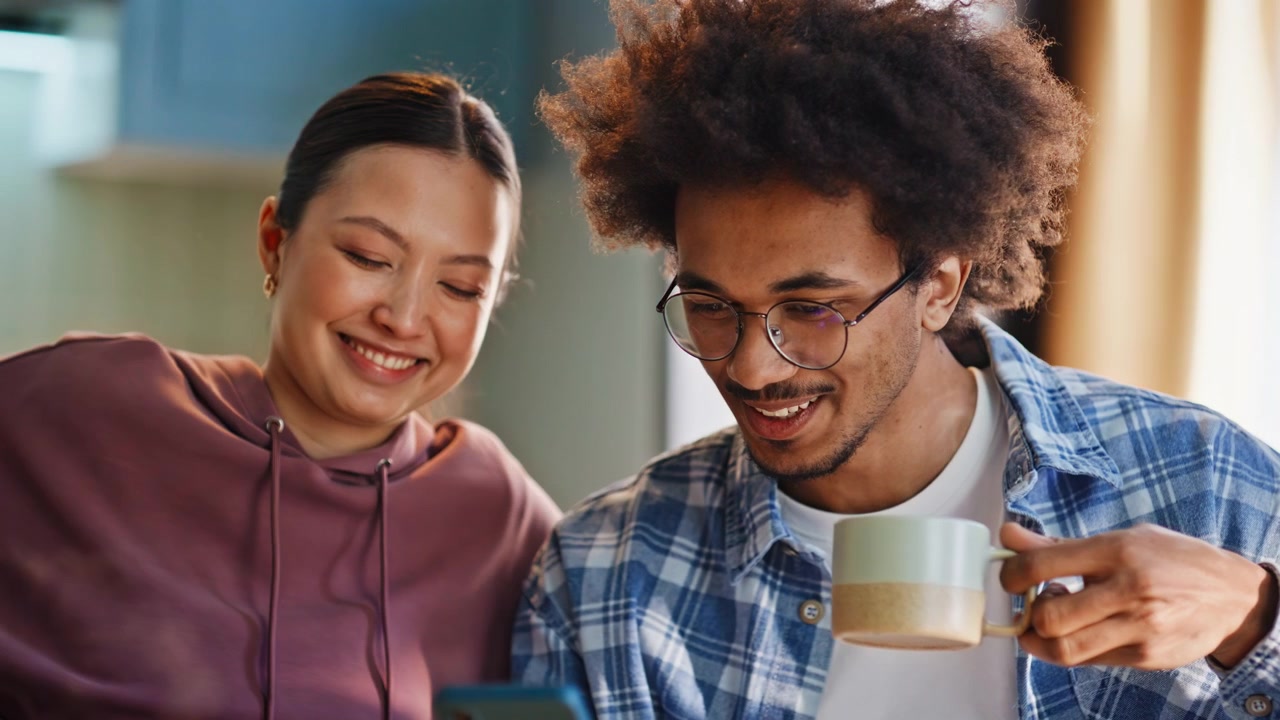 Multiethnic couple browsing smartphone over morning coffee in home kitchen