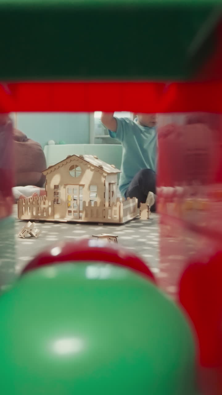 초점을 맞춘 금발 소년은 다채로운 블록으로 벽을 건설합니다. 유아는 바닥에 앉아 어져있는 장난감으로 놀고 큐브의 건설에서 클리어런스를 통해 볼 수 있습니다.