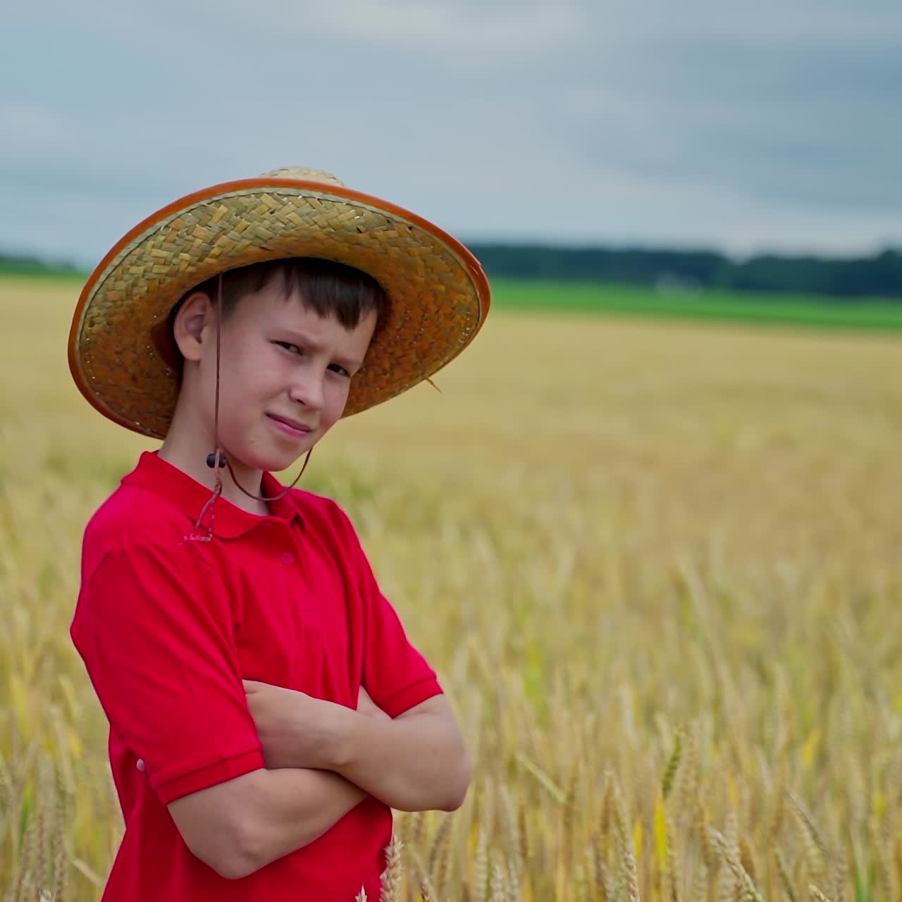 Teenager in straw hat among nature. Portrait of a pensive boy standing on a wheat field in summer. Serious child on the background of yellow farmland.