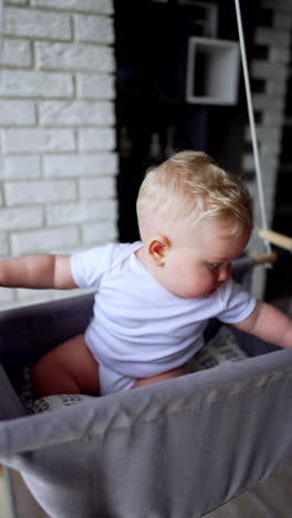 caucasian baby with plump cheeks sits in the swing indoors. happy kid having fun. vertical video.